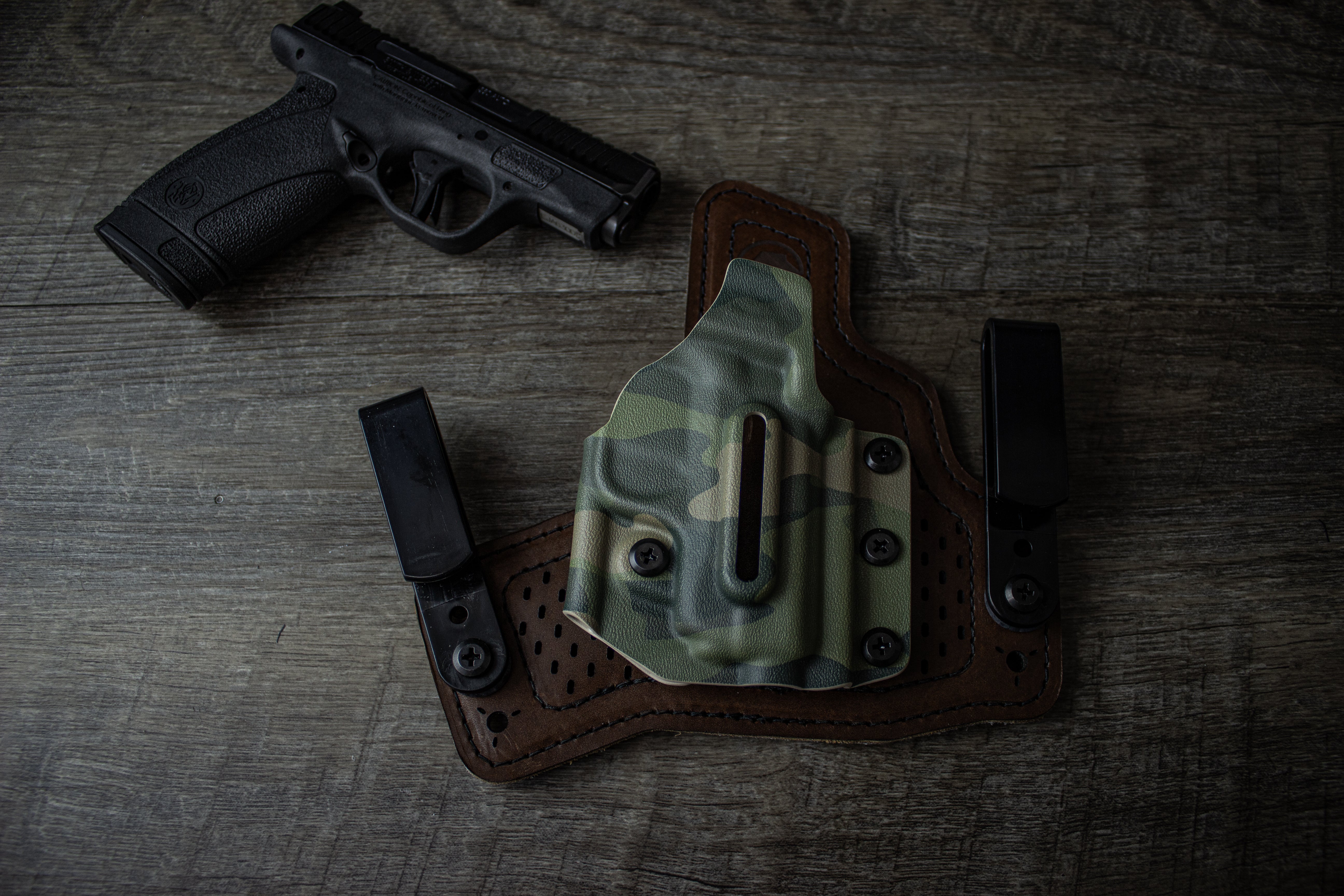 Black Arch Protos-M holster in Multicam Tropic Kydex with dual polymer clips and brown leather backer, shown beside a Smith & Wesson Bodyguard 2.0 pistol on wood surface.